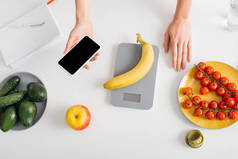 举着智能手机的女孩在厨房桌子上称香蕉的俯视图，热量计算饮食