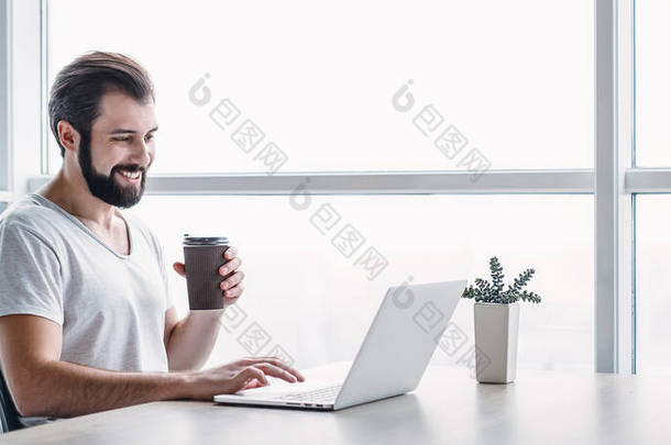 带着笔记本电脑和咖啡坐在办公室里的面带微笑的男人的肖像的网站首页。侧视图