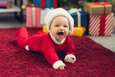 笑小婴儿穿着圣诞老人西装躺在圣诞树前的红地毯和礼物