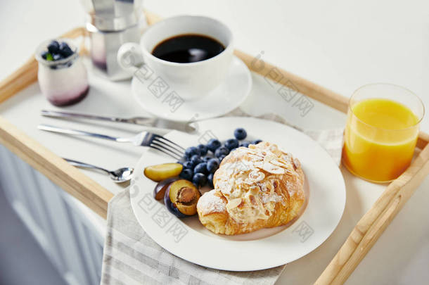 在白色桌面上的<strong>木托盘</strong>上, 用新鲜的蓝莓和李子片、杯汁、杯咖啡和酸奶来接近牛角面包