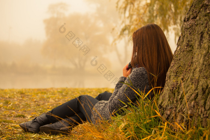 孤独的女子在一个有雾的秋日近水树下休息。孤独的女子在秋天享受自然景观。秋季的一天。女孩坐在草地水平图像.