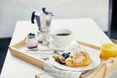 在白色桌面上的木托盘上, 用新鲜的蓝莓和李子片、杯汁、杯咖啡和酸奶来接近牛角面包