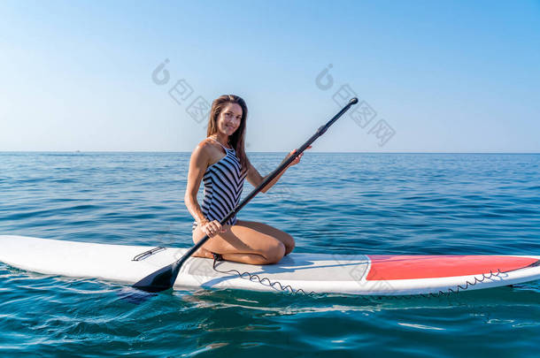 阳光明媚的夏日,一个身材苗条的女孩在海上滑板上滑行.穿着条纹泳衣，在酒吧里。风暴在海上的夏季活动