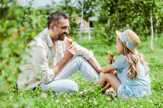 选择的焦点欢快的父亲看着孩子在草帽与冰淇淋圆锥坐在草地上 