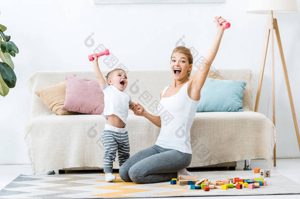 笑的母亲和幼儿男孩拿着哑铃在举起手, 看着镜头在客厅的地毯上