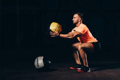 英俊的运动运动员做蹲在黑暗的健身房医学球
