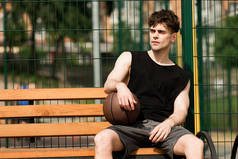 在阳光明媚的一天，在篮球场上，有球坐在木凳上的严肃的篮球运动员 