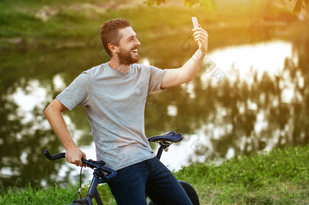 穿着灰色T恤和蓝色牛仔裤骑着自行车的英俊年轻人- -在湖边绿树成荫的小巷里，电话里挂着一张自拍照片 