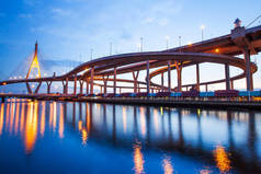黄昏时分，高低角的公路交接处和悬索桥景观映照下，朝弗拉亚河的水面反射出发亮的灯光。泰国普密蓬大桥。长期接触.