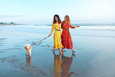 夏天。带着狗在海滩上的女人马西波希米亚服装时尚少女们带着宠物在热带海洋附近的爱犬海岸散步。湿沙上的轮廓反射和脚印.