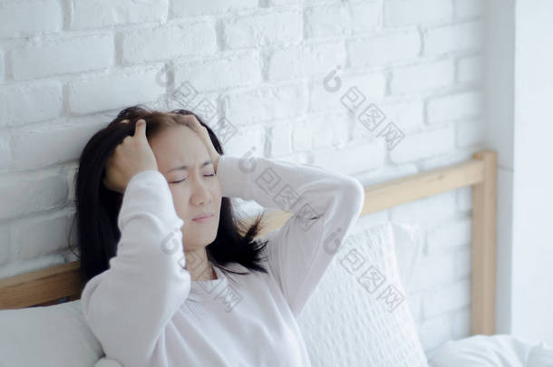 亚洲妇女头痛严重。女士早上醒来时偏头痛。失眠在醒来时会导致头痛。坐在压力床上的年轻女孩。在她的卧室里她病了.