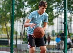 一个穿着绿色T恤的可爱男孩在城市的操场上打篮球。活跃的青少年享受户外游戏与橙色球。爱好、积极的生活方式、孩子们的运动.