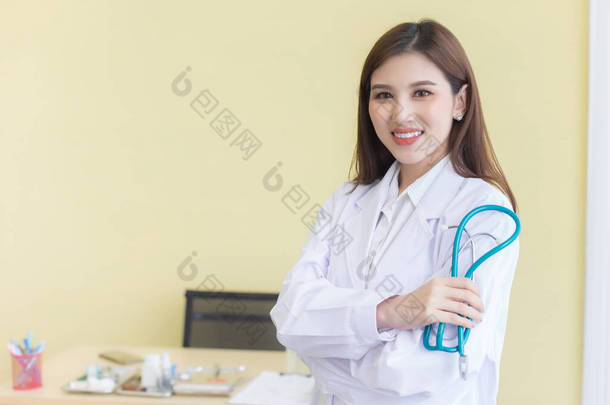 年轻美丽的亚洲女医生抱着<strong>胳膊</strong>高高兴兴地站在医院里。穿着白袍和听诊器
