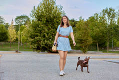 穿着蓝色衣服的年轻漂亮的、快乐的黑发女孩和她的狗在一个城市公园里散步。法老的狗在外面散步 . 
