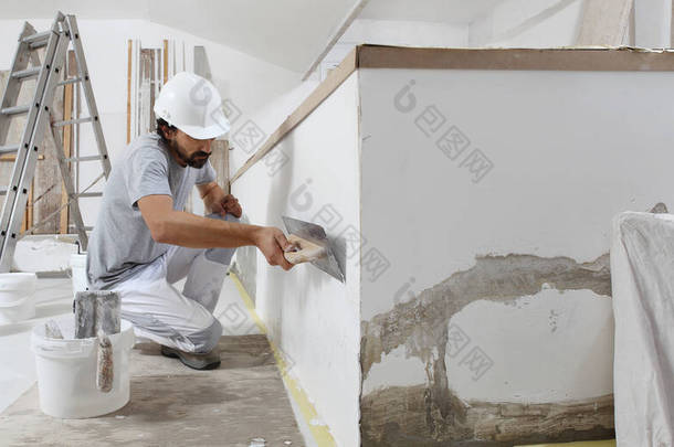 建筑工人在工作时，把灰泥从桶中取出，用铁钳固定在墙上，在房屋的建筑工地上戴上头盔