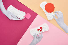 剪纸白手与白色卫生巾附近的空卡和血滴在紫色，粉红色和米色背景的视图