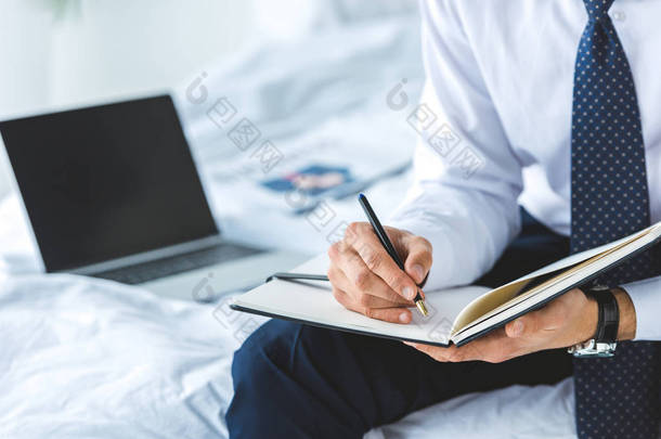 用笔记本电脑<strong>坐在床上</strong>写日记的商人的裁剪视图