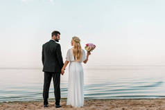 一对夫妇站在沙滩上的婚礼花束和看着对方的情侣后视图