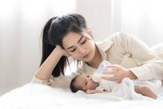 年轻的亚洲母亲抱着一个可爱的新生儿，在白色的卧室里喂奶瓶。母亲和儿童概念