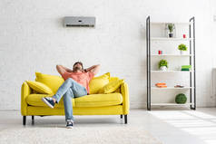 在宽敞的公寓里，一个年轻人坐在空调机下的黄色沙发上