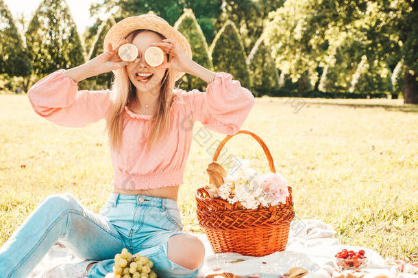 年轻美丽的嬉皮士女人穿着时髦的夏季牛仔裤，粉色T<strong>恤</strong>和帽子。外面野餐的女人正面的模特坐在草地上，吃着水果和奶酪，手里拿着柠檬 