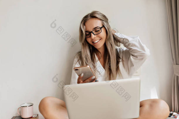 现代成功的女孩，金发碧眼，头戴眼镜，带着笔记本电脑和智能手机，在阳光下与外界隔绝的白色背景下工作