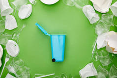 蓝色回收站在绿色皱褶的塑料杯、叉子、盘子和纸板容器之间的顶视图
