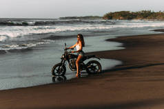美丽的单身女孩在海滩上骑摩托车与日落相遇。在海边骑自行车的时髦女人.骑摩托车旅行。去巴厘岛的旅行热带海滩上的一辆定制摩托车.复制空间