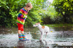 孩子和狗在秋天公园的雨中玩耍.小孩走狗。下雨天,小男孩在泥泞的水坑里跳跃.雨靴和夹克，户外穿着。儿童防水鞋和外套.