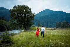 美丽幸福的情侣的肖像牵着手, 沿着雏菊草甸走在山上的河边, 覆盖着绿树.