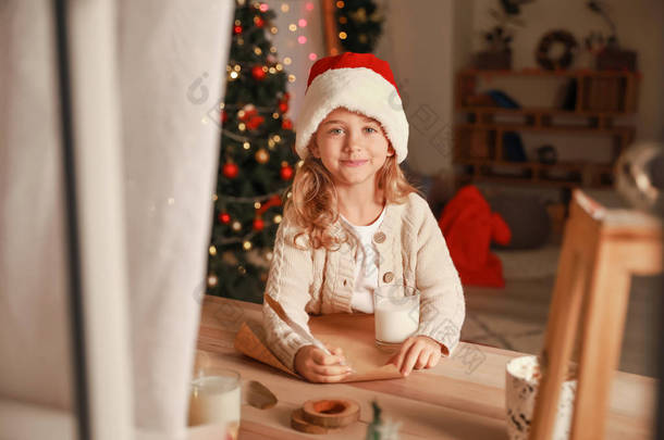 可爱的小女孩写信给圣诞老人在圣诞节前夕在家里。通过窗口查看
