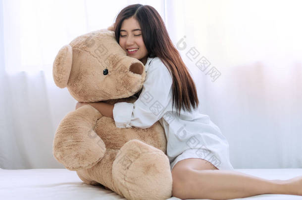 迷人的笑着快乐的年轻女子抱着她的大泰迪熊<strong>坐在</strong>家里的白色<strong>床上</strong>。可爱的女孩在卧室里欢快地拥抱着棕色的泰迪熊.