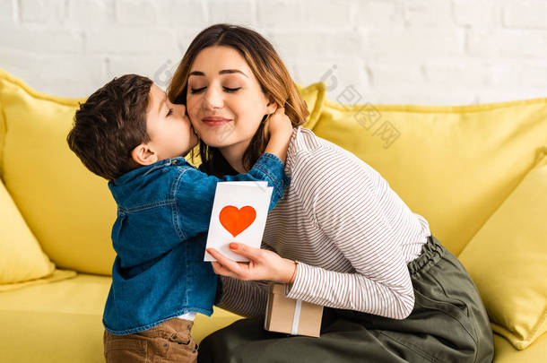 可爱的男孩吻着快乐的妈妈，手里拿着礼品盒和印有心形符号的母亲节贺卡