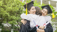 两个亚洲女人在毕业礼服和帽子上微笑和感到快乐, 和父母在手臂拥抱中站在一起