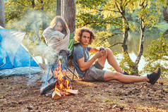年轻的夫妇在阳光明媚的日子里, 在森林里露营的篝火旁取暖。.