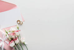 在灰色背景上, 带粉红色信封、鲜花、包裹的礼物和金色结婚戒指的空卡的顶部视图