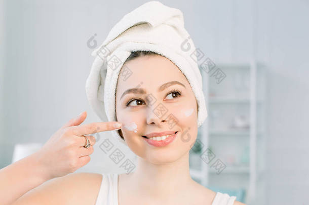 在她的头上有条毛巾的快乐的年轻女孩在她的脸上涂抹一个<strong>洁面面</strong>具