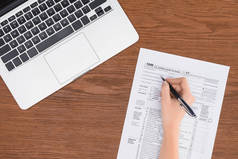 裁剪视图的妇女填写税表在工作场所与笔记本电脑