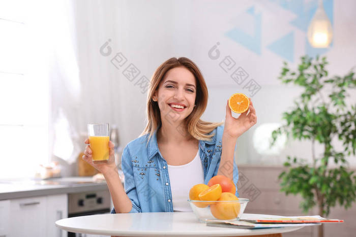 快乐的年轻女子与一杯果汁和橘子在厨房的桌子。健康饮食