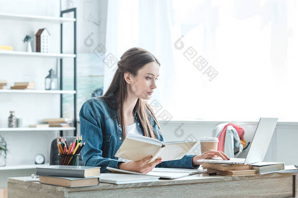 重点青少年女孩拿着书和使用笔记本电脑, 而在家学习