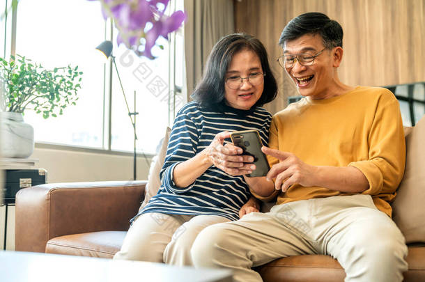 快乐的老亚洲情人夫妇手握智能手机，看着手机屏幕轻松地<strong>坐在沙发上</strong>，笑着年长的祖父母家人拥抱生活方式