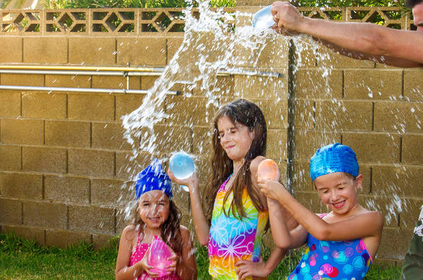 父亲的<strong>胳膊</strong>在他的三个女儿的头上打碎了一个蓝色的水气球。女孩们穿着泳衣, 其中两人有游泳帽上.