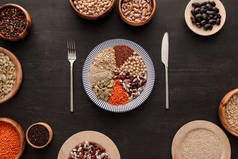 在深色木质表面，带各种生豆类、谷类和碗的带条纹盘子附近的刀叉顶视图
