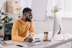 英俊的非洲裔美国设计师在橙色毛衣绘图在笔记本电脑和看在办公室的计算机