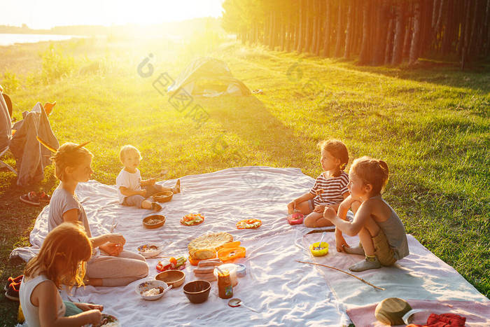 孩子们在草坪上野餐，坐在毛毯上吃饭。柔和的夕阳西下.