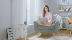 在舒适的客厅里，年轻布鲁内特的肖像在家里的笔记本电脑上工作。穿着粉红汗衫的漂亮女孩，消磨时光.