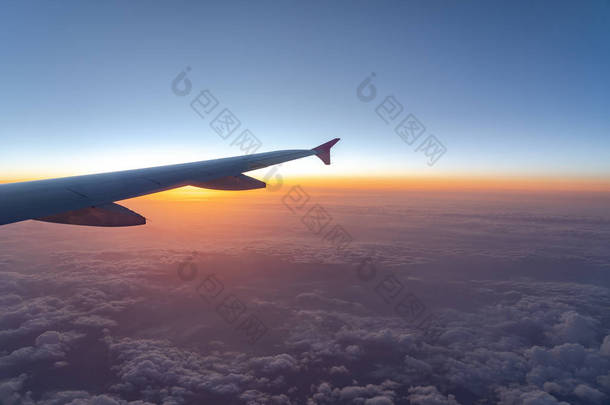 在空中，从飞机窗口看，在太阳升起时，有深蓝色天空地平线和<strong>云背景</strong>的飞机机翼剪影