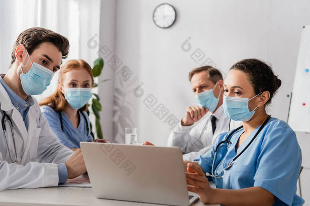 戴着医疗面具的多文化<strong>医院</strong>工作人员坐在<strong>医院</strong>的工作场所，在模糊的背景下看着笔记本电脑