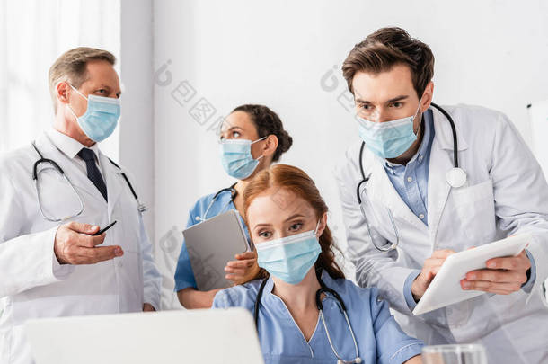 戴着医疗面罩的护士在<strong>医院</strong>多<strong>文化</strong>同事身边看着笔记本电脑，前景暗淡