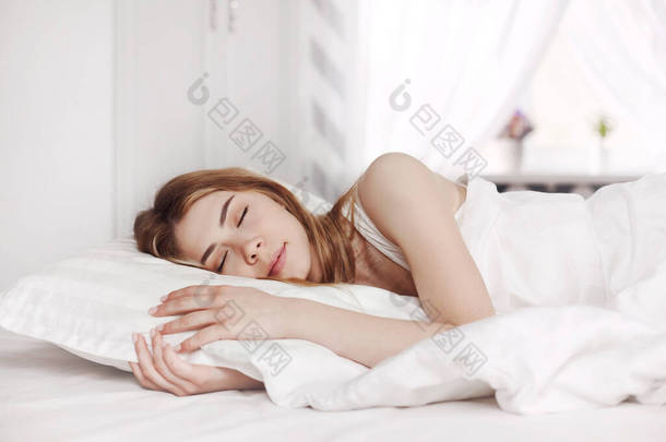 早上，一个穿着白色睡衣的年轻貌美的<strong>女孩</strong>睡在她家明亮<strong>卧室</strong>的床上。侧视图、文字空间